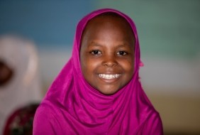 A young girl wearing a pink headscarf looks at the camera and smiles.